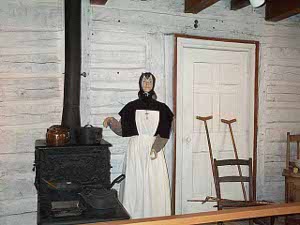 The Exhibit shows a nun wearing a black cowl and a white dress standing next to a stove; a pair of crutches rest near the adjacent door.