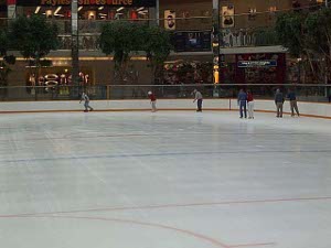 Severalskaters circle the large rink (marked for hockey) in the West Edmonton Mall.  Stores can be seen on two levels in the background.