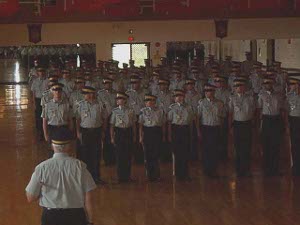The large drill hall has a polished floor; the trainees, attired in summer uniforms, in various sizes and shapes, are performing a dress right command.