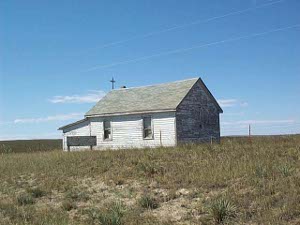 A wooden frame building with space for two windows on one side, and a cross on the roof, surrounded by wild grasses, was formerly a prairie church