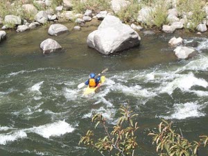 Wearing a blue helmet and orange vest, a man in a yellow kayak negotiates the river rapids, with large boulders along the banks.