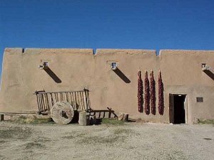 A plain stucco wall fronts the adobe, with a wooden-wheeled cart positioned in front.  Large gutters drain the rainwater from the roof, while four giant strings of chilies are positioned to the left of the door