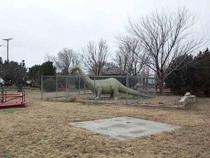 The dinosaur is protected by a cyclone fence in a small park and playground