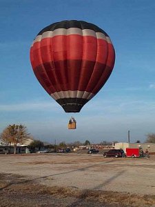 The balloon, colored two shades of red with white and blue stripes, from which a basket with three people is suspended, hovers a little above the cement take-off location