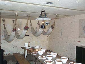 Dishes of food are set out along a long trestle table; overhead are slung hammocks from ceiling hooks indicating the room doubled as a bedroom