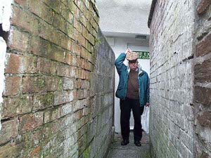 The alley is barely wider than Bob, who stands holding his hat and preparing to squeeze his guts through the alley