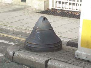 A cast iron bell shape sits on a traffic island near the sidewalk