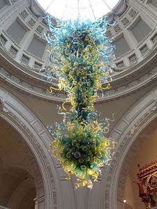 The hanging glass ornament is perhaps six feet high and three feet in diameter at the widest, suspended from the domed roof of the V&A.  The glass is colored green and yellow with blue glass near the top, all in thousands of tiny, wildly curving tendrils of glass.
