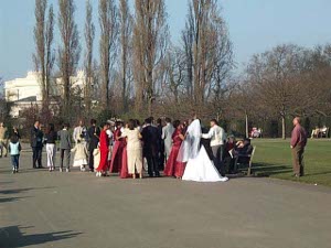 About twenty wedding participants cluster around the edge of one of the gravel paths in Regent's Park.  The bride wears white, with a veil descending to her hips, the bridesmaids in bright red