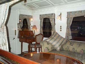 The inside of the parlor car has a large sofa and an even larger table, upon which food and beverages might be placed.  A desk in chair is in one corner, the large windows are curtained, and lights are mounted on the white walls.