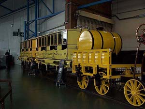 A yellow wagon has a large water tank, and is followed by two yellow and black passenger cars.
