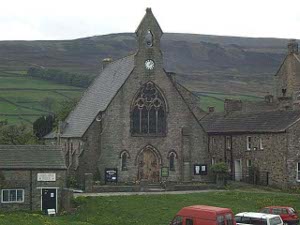 The dark stone church has a steep roof; in the background are walled green sheep pastures, and above, atop the hills, the barren moorland.  The gray day enhances the drearer dull colors, brown, and green