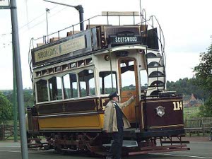 The double-decker tram car is painted a bright maroon and orange, with vintage advertising, and a properly dressed conductor