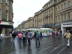 The paving stones a little wet from a recent rain, vendors have set up market stalls covered with transparent green plastic to sell produce in a market located in the Newcastle pedestrian era, flanked by four-story brick buildings.