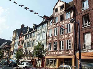 A row of four story timbered buildings provides a beautiful facade to a public square in Rouen.  Vehicles line the street, trees are planted, and the first story is given over to shops and businesses