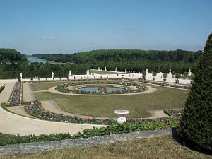 The immense formal garden with a circular pond and fountain in the center leads to long formal walks lined with statues and, in the distance, a beautiful man-made lake surrounded by forests, all part of the vast lands of Versailles