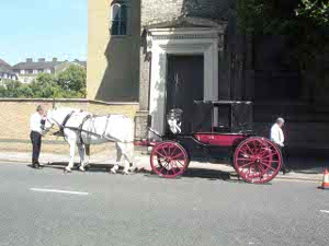 An old black and red carriage, pulled by two white horses, is tended along the side of Albany Street