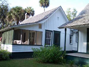 The white frame home has a screened in porch and green trim, with palm trees in the background