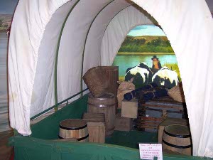 A reconstruction of a covered wagon has green wooden frame and iron hoops, with a white canvas cover.  Inside are tightly closed wooden crates and barrels.