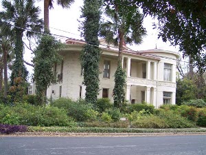 A cream colored two-story mansion with stone coigns, a circular wing, hidden behind ivy-covered palm trees