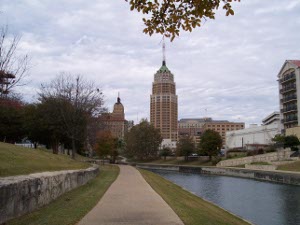 Cloudy sky, downtown buildings in the center, including a stepped skyscraper with a conical green copper roof, and the San Antonio River in the foreground
