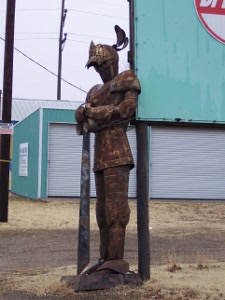 In front of a commercial garage stands a rusted iron knight, sculpted out of sheet metal.
