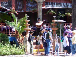 People stroll along the sunny San Antonio River Walk in front of a restaurant with musicians in Mexican attire.