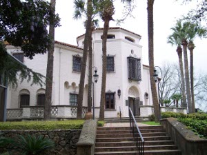 The mansion sits on top a hill, built in Mediterranean style, with iron railings over the windows and a red tile roof, surrounded by palm trees.