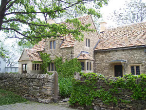 The beautiful tan stone Cotswold cottage sits behind a low stone wall, covered with ivy.