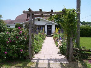 There is a wooden trellis leading up a garden path to the front door of the white frame building which is Garden's Gate Restaurant