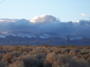 In the foreground, desert brush, topped by a line of power poles.  In the background, the Sierra Nevada mountain range, viewed from the East, snow-capped, topped by fleecy grey and white clouds, and a light blue winter sky above all.