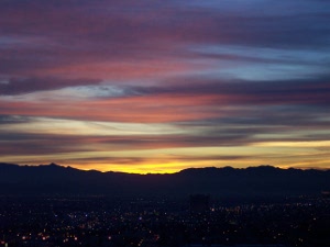 The light blue sky is streaked with scarlet and purple, and a bright yellow light at the horizon, below which the mountains seem black, with sparkles of Las Vegas night lights.