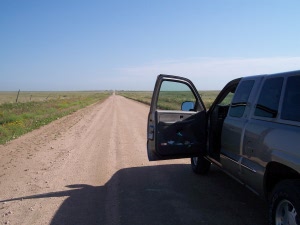 A straight flat dirt road flanked by empty green pastures stretches as far as the eye can see, next to the open door of our truck.