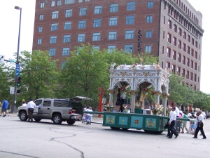 An elaborate white and gold shrine supported on six pillars separated by curved arches over a decorated blue base is pulled through the streets of Omaha by a pickup truck and escorted by men in dark pants and white shirts.