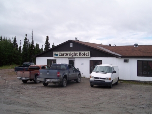 Two trucks and a van are parked in the dirt in front of the Cartwright Hotel, white siding with a brown shingle roof.