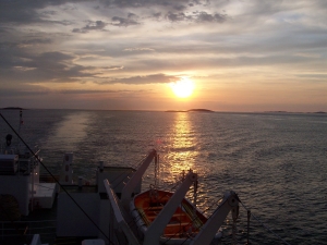 Sunrise from the ferry boat; the lifeboat hangs from davits, the sun casts a reflection across the sheltered inland waters, parallel to the ferry's glassy wake, with sun rays in the cloudy sky shading from orange to gray.