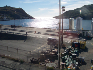 The afternoon sun glistens on the water leading out of St. John's protected harbor to the sea.  On the oil dock can be seen tanks and drums with different colored lids filled with materials for offshore oil well construction.