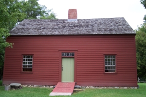 The salt-box house is painted colonial red, with one window on each side of a plain center door, and a central fireplace.