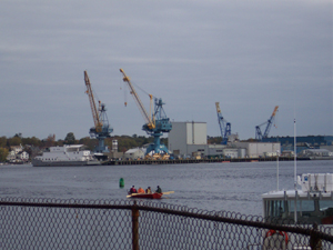 A red rowboat with four men crosses the harbour towards a large 
waterfront facility with many cargo cranes