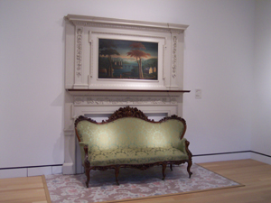 A painting is mounted in a panel above a fireplace in this white-
walled gallery.  In front sits a lovely carved sofa upholstered in green brocade.