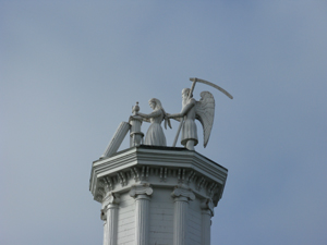 The sculpture is from a single redwood tree, painted white, on top of a high tower on the former Masonic Lodge.  The maiden stands holding a book at a pedestal, while behind her, holding her hair, stands a male figure with wings, carrying a large wooden scythe.