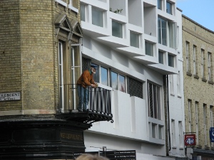 The mannikin has a white beard, blue hat, orange sweater and blue trousers, and is holding on to the rail of the balcony.