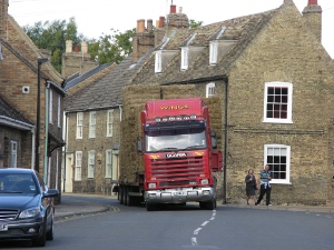 The red semi-trailer truck has hay piled to a height of perhaps 15 feet or more; it is backing up around a curve in a narrow street lined with houses, parked cars, and pedestrians.