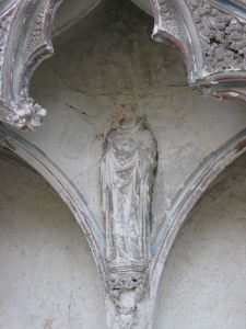 In a corner surrounded by curving arches and filigree, a statue of a saint has the head broken off at the neck