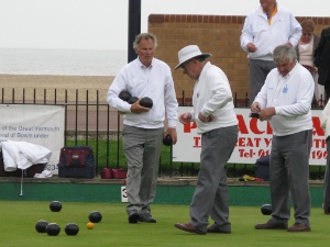 Three grey-haired lawn bowlers clad in grey trousers and white sweaters gather up the balls.  The bowling green is next to the sandy beach.