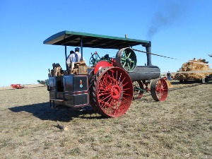 An old steam tractor has bright red wheels and a green roof over a gray boiler and steam engine.  A power takeoff wheel provides power to farm equipment by a belt drive.
