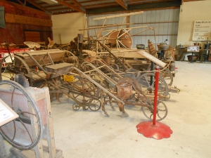 Old plows and farm equipment are displayed on the floor of a large open building