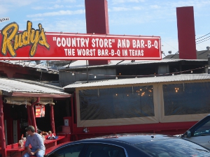 The sign reads Rudy's Country Store and Bar-B-Q and the restaurant is a simple wooden building.