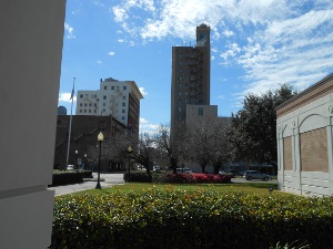 The sky islight blue with patches of fluffy white clouds; the tan and ochre sides of the museum building are in the foreground, and over a hedge of greenery rises two high rise buildings, the one topped with a clock tower.