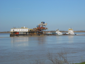 The barge has several tugs pushing it upstream; it is equipped with cranes for some kind of river maintenance operations